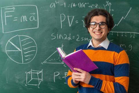 smiling-young-geometry-teacher-wearing-glasses-standing-front-chalkboard-classroom-holding-open-book-looking-front-min